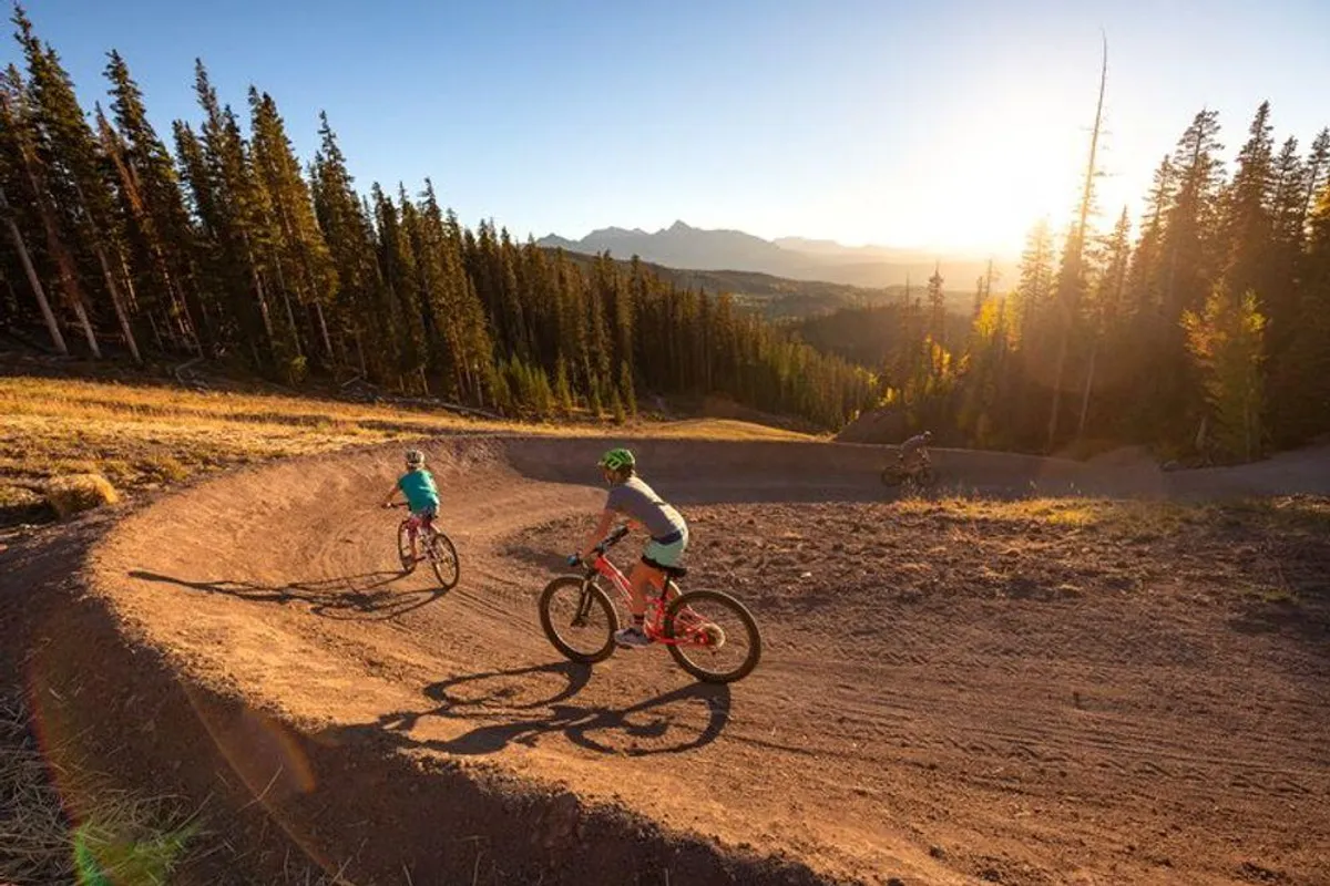 An action-filled exterior image featuring mountain bike riders navigating a curved dirt trail through dense pine forest as the sun sets. The energy of the riders and the vibrant setting evoke the adventurous spirit of Telluride’s mountain trails.
