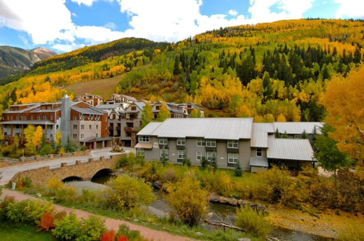 A picturesque riverside setting with colorful autumn foliage, a peaceful river crossed by a bridge, and modern buildings with a mountain backdrop. The scenery is tranquil and inviting, suggesting outdoor exploration and family-friendly adventures.