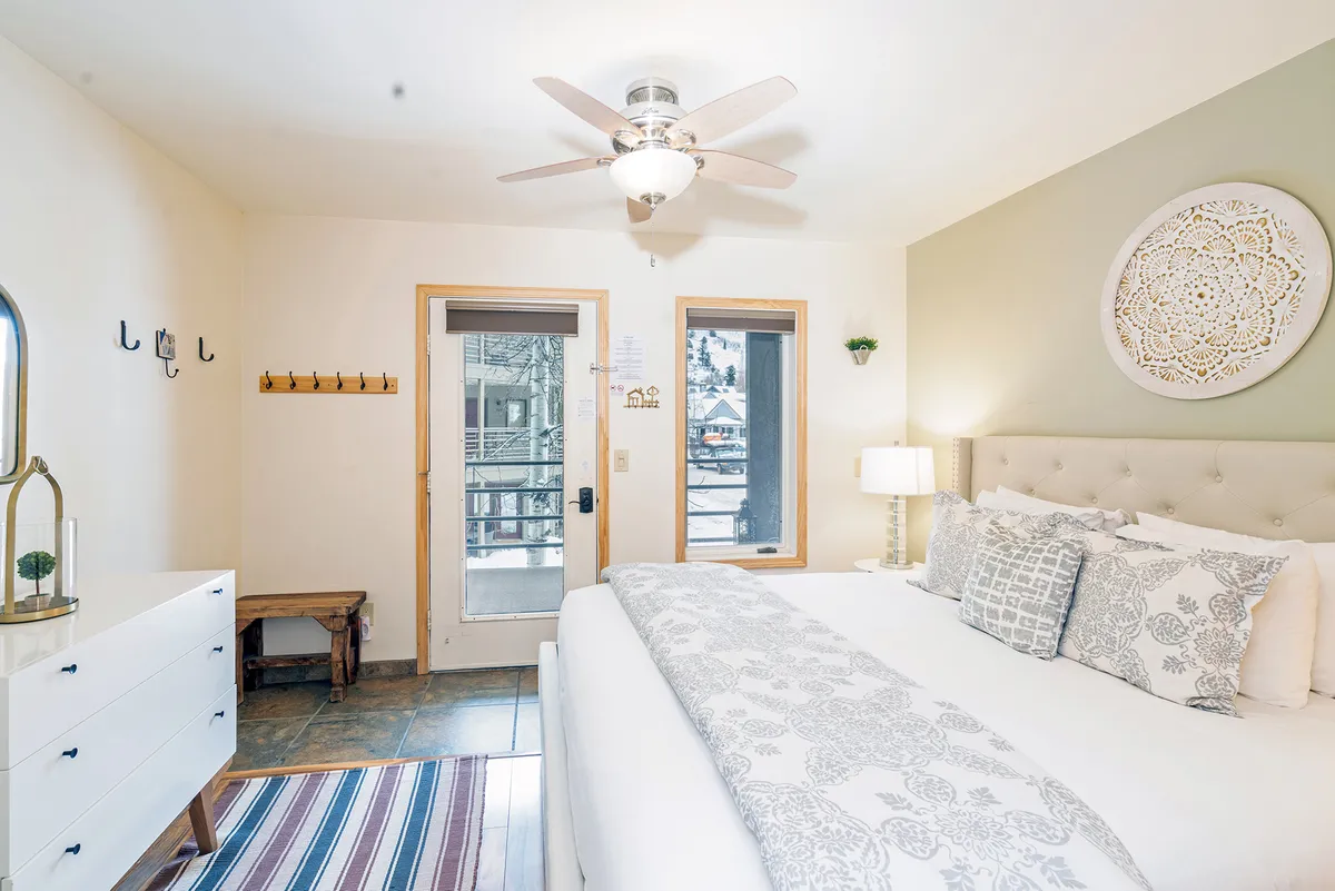 A tranquil bedroom featuring a neatly made bed adorned with decorative pillows, a window that lets in natural light with a view outside, and a dresser topped with decorative items. The space feels calm and inviting, designed for restful moments and relaxation.