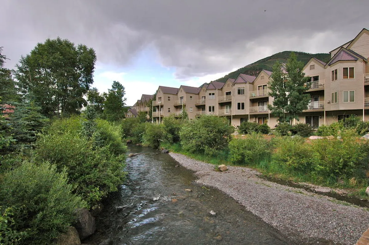 This exterior shot captures a peaceful stream winding beside lush greenery and residential buildings, framed by mountain vistas. The scene exudes tranquility, with the waterway and walking paths hinting at direct outdoor access from the property.