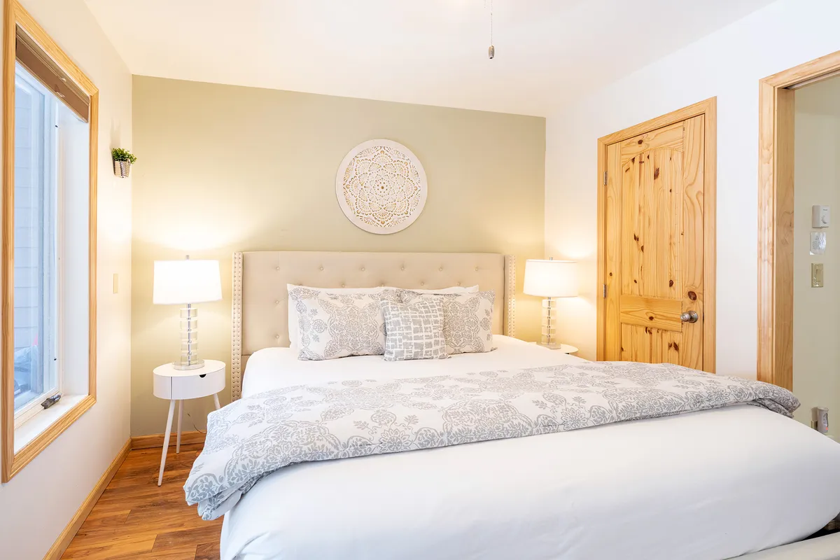The image showcases a serene bedroom with a plush king-sized bed adorned with decorative pillows, framed wall art, cozy nightstands with softly glowing lamps, and a large window that fills the space with natural light, offering a glimpse of the peaceful hillside beyond.