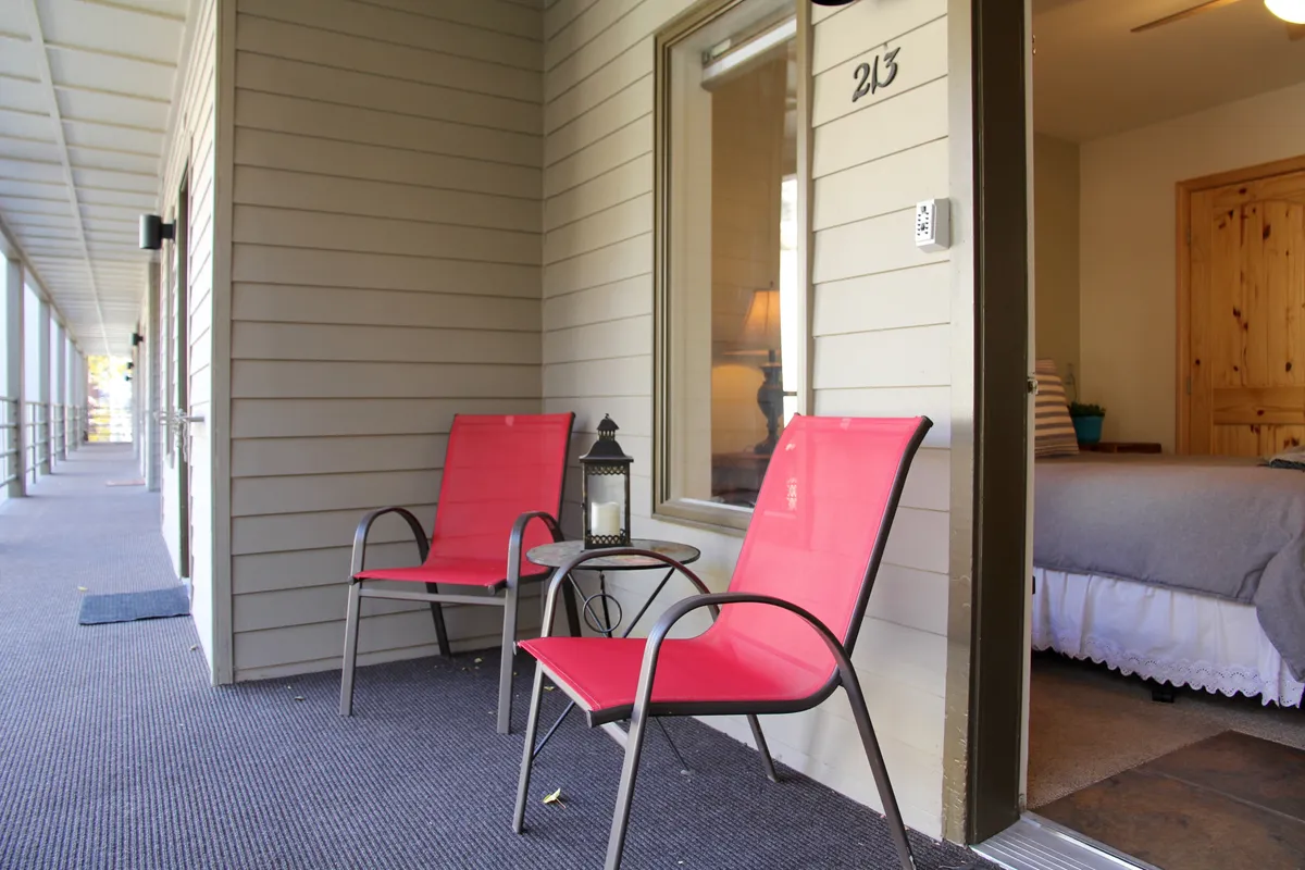 A charming exterior setting featuring two red chairs and a small table on a porch, with a lantern nearby and a welcoming doorway. The scene is bathed in warm light, suggesting a relaxing spot for morning coffee, reading, or enjoying quiet conversation while soaking in the outside atmosphere.