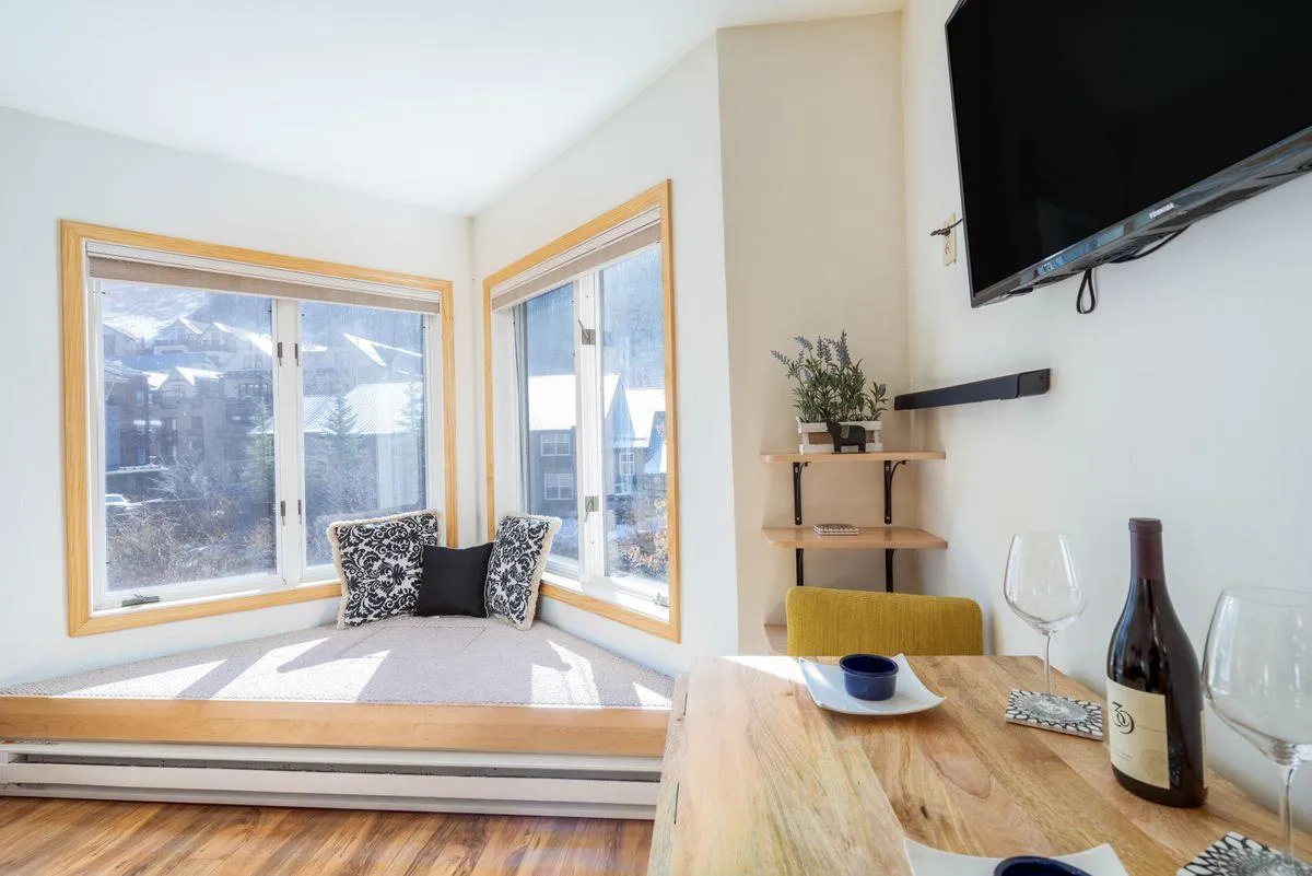 The image highlights a cozy bay window seating area, next to a dining table set with wine, and a shelf adorned with green plants. Sunlight pours in, illuminating the intimate dining and relaxation space, while the view through the bay window hints at mountain vistas.