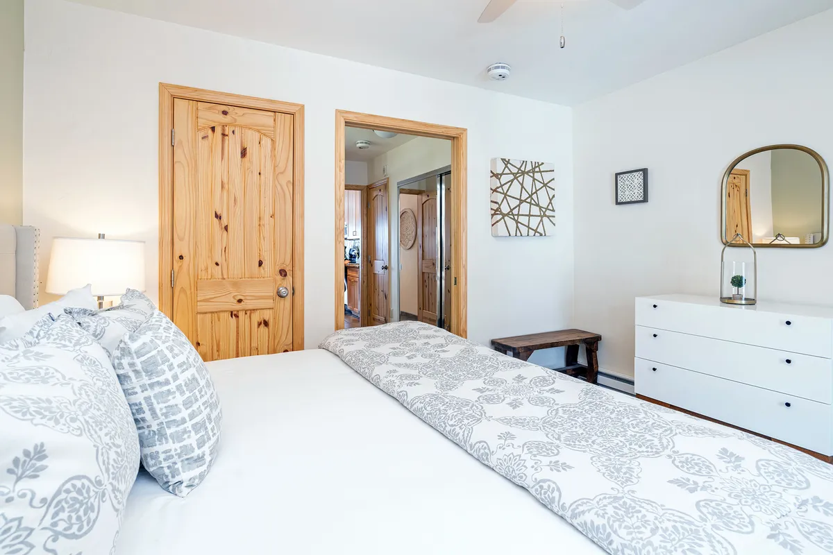 A serene bedroom scene with a patterned bedspread, a wooden door, a dresser, and a large mirror. The soft, calming mood is accentuated by thoughtful details and a sense of ample storage.