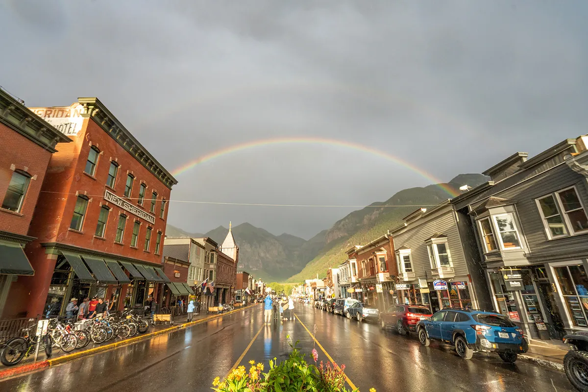 A lively Telluride street scene featuring historic buildings, a vibrant rainbow arching across a wet street, bicycles, and people strolling along the sidewalks. The atmosphere is cheerful, suggesting a walkable, welcoming small-town vibe ideal for families.