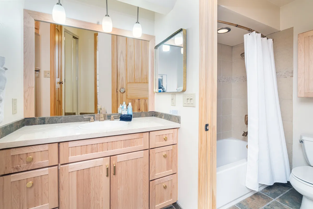 This image features a contemporary bathroom suite with a sleek vanity and sink, expansive mirrors, a modern shower area, toilet, and stylish wooden cabinetry. The space is clean, well-lit, and organized, with quality towels visible, creating a serene and inviting atmosphere for both quick routines and relaxed bathing.