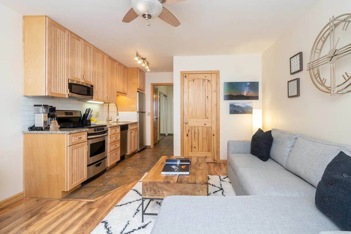 This image presents an inviting open-concept living space, where the kitchen area with visible appliances and cabinetry seamlessly connects to a comfortable sofa and coffee table. The scene includes wall art and a clock, reinforcing the communal and homely ambiance. The layout clearly showcases the kitchen’s proximity to the living area, ideal for gatherings and shared moments.