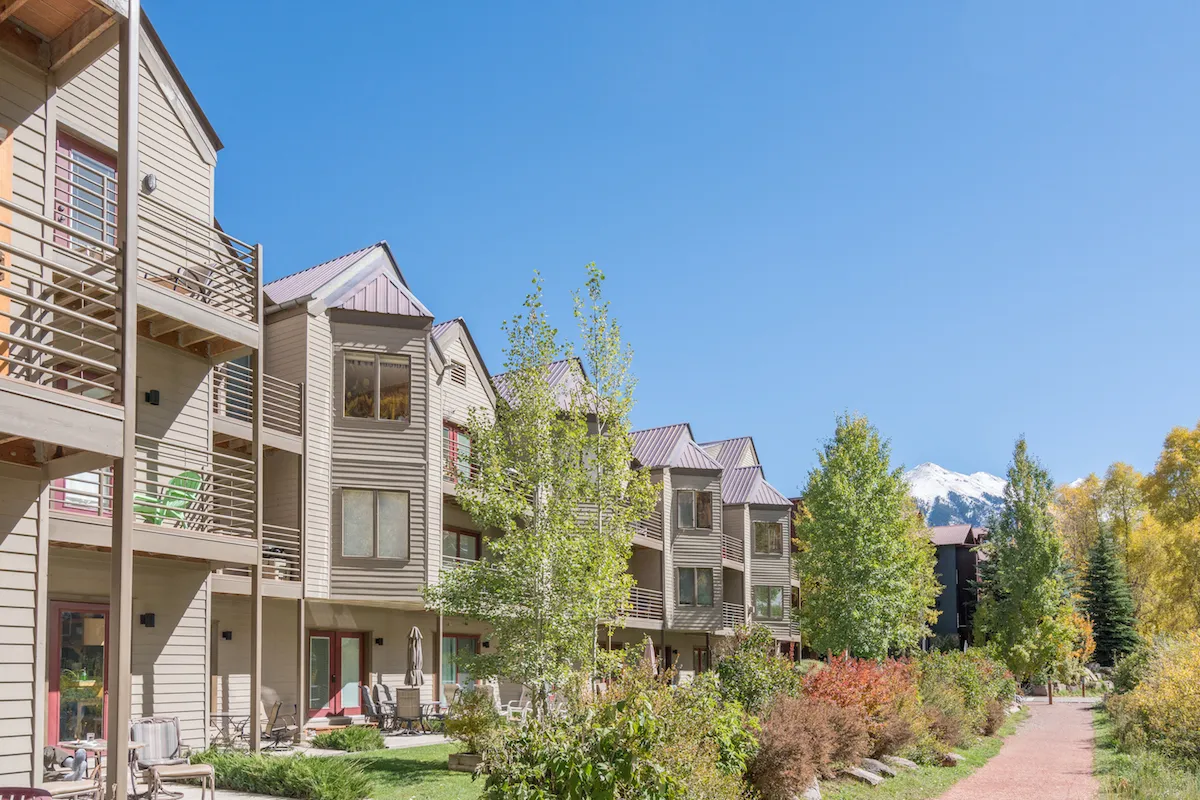 This image showcases the exterior of the property, with balconies overlooking green trees and a stunning mountain backdrop. The setting is tranquil, and the path lined with greenery suggests easy walkability in a scenic environment, aligning with downtown Telluride's outdoor appeal.